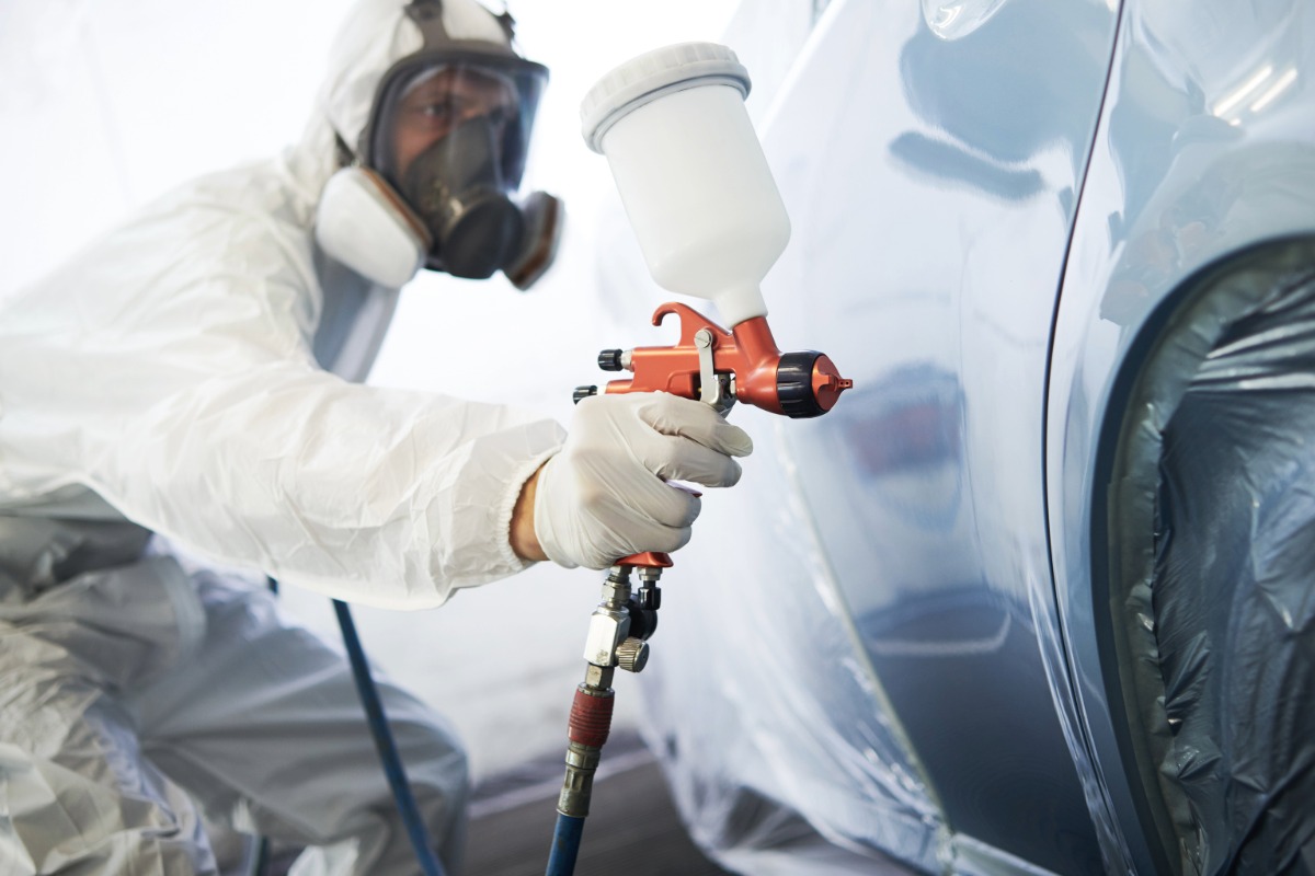 Technician in protective gear using a spray gun for collision repair on a vehicle in a modern auto body shop.