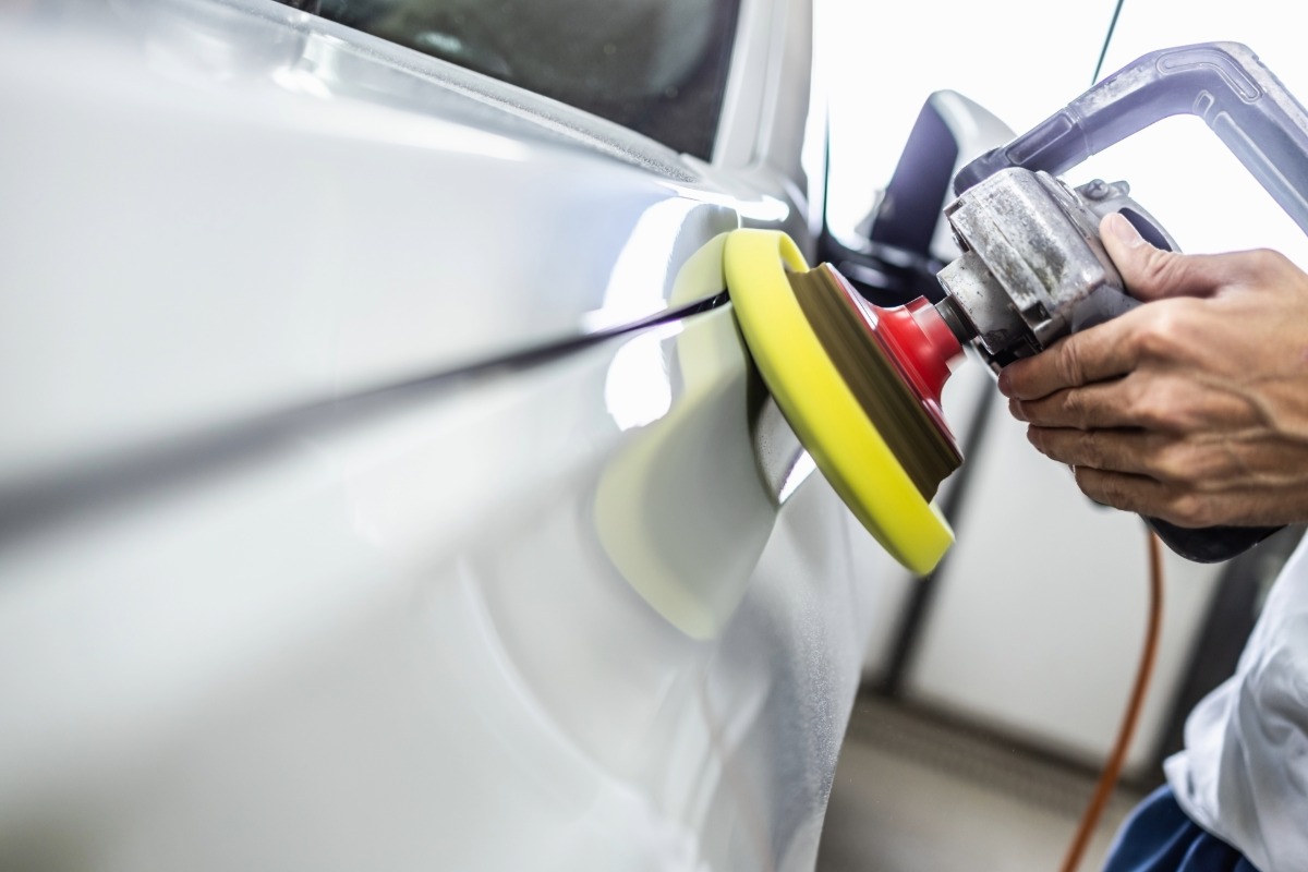 Automotive technician polishing a vehicle's surface with a rotary buffer in a collision repair facility.